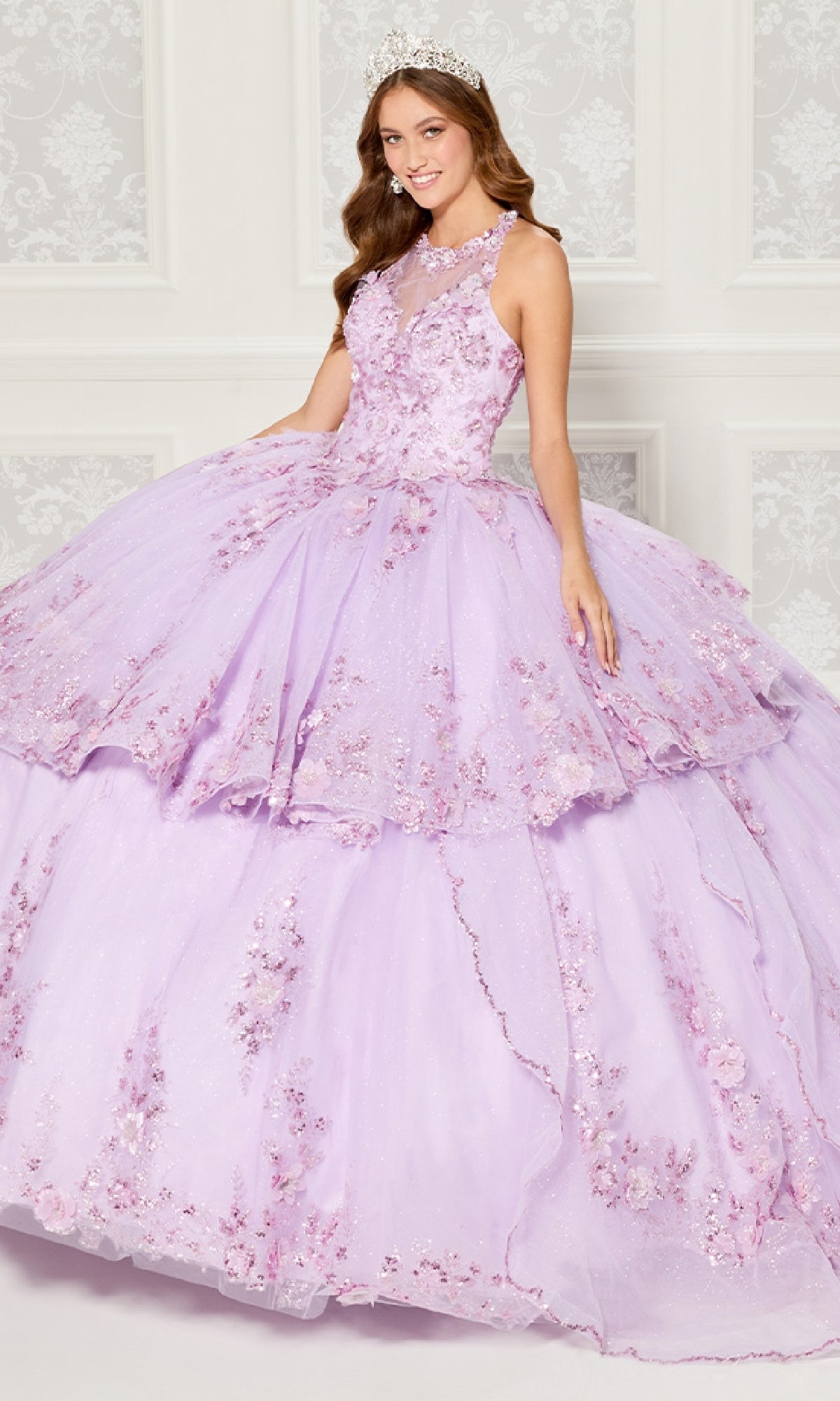 A young woman in the Quinceanera Dress PR30082 By Princesa, featuring light purple fabric and delicate floral embroidery, poses with a tiara in front of a white patterned wall.